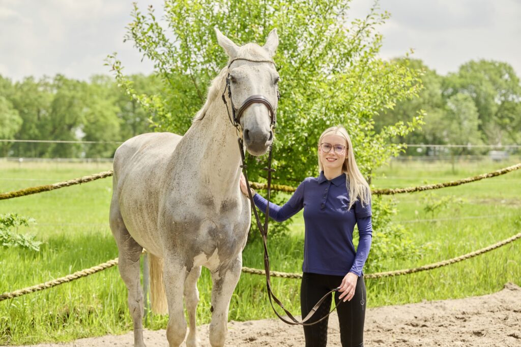 12-Tierarzthelferin-mit-Pferd-Tierarztpraxis-Friedeburg Frau steht mit weißem Pferd vor grüner Wiese